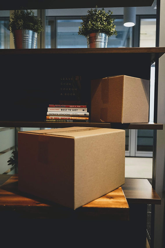 Cardboard box full of books ready to ship resting on desk. Books in background over the top of the box on a wooden shelf.