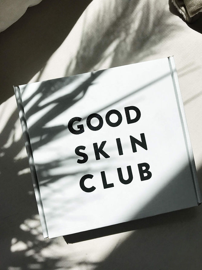 White shipping box with brand logo "Good Skin Club" in black, capital lettering. A slight, branch-like shadow is cast over the white packaging.